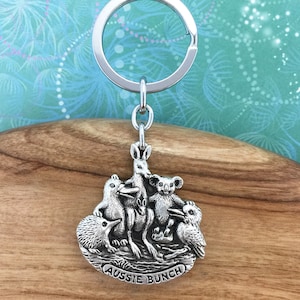 May include: Silver-colored keychain featuring an Australian animal design. The keychain has a ring at the top and a charm with a kangaroo, koala, echidna, owl, and other animals. The text "AUSSIE BUNCH" is at the bottom of the charm.