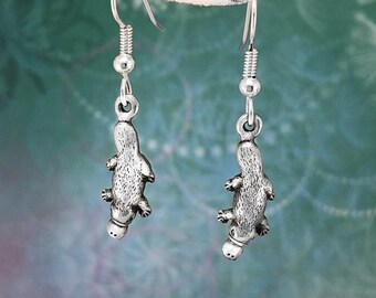 May include: Silver-coloured earrings, each with a detailed platypus charm. The earrings hang from a simple hook with a coiled spring detail. The background is a soft green with floral accents, highlighting the unique shape of the aquatic mammal.