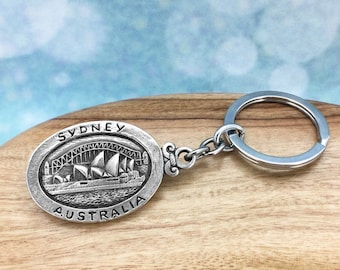 May include: Silver-toned keychain featuring a detailed depiction of the Sydney Opera House and Harbour Bridge. The oval-shaped charm reads "Sydney Australia" in a raised font.