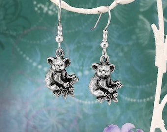 May include: A pair of silver dangle earrings featuring a koala bear charm sitting on a branch with flowers. The earrings are hanging from a white branch.