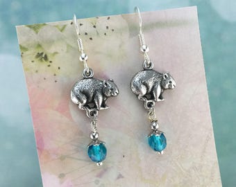 May include: Silver dangle earrings featuring a small, detailed wombat charm and a dangling turquoise crystal bead.
