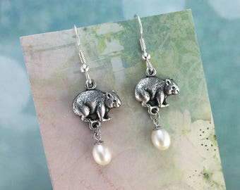 May include: Silver dangle earrings featuring a small, detailed wombat charm and a single white pearl hanging below.