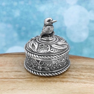 May include: A silver trinket box with a detailed design of leaves and vines. The lid features a sculpted bird with a raised beak.