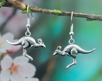 May include: A pair of silver kangaroo earrings. Each earring has a detailed kangaroo pendant suspended from a silver hook. The kangaroos are depicted in a leaping posture, with their tails extended. The background is a soft blur of green and white.