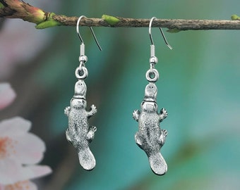 May include: A pair of silver earrings featuring a platypus design. The earrings are hanging from a branch.