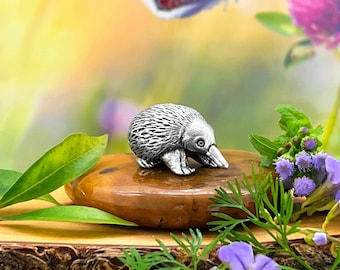 May include: A small, silver echidna figurine sitting on a brown rock with green leaves and purple flowers in the background.