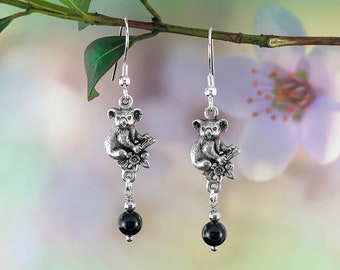 May include: Silver koala bear drop earrings. Each earring has a detailed koala figure on a branch with flowers, suspended below a black, round bead. The earrings hang from a simple hook.