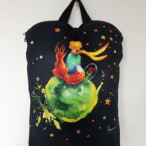 Pode incluir: Sacola de lona preta com abertura em forma de coração. A sacola apresenta uma ilustração colorida do Pequeno Príncipe, uma raposa e uma rosa em um planeta verde com estrelas no fundo. O texto "Rasmus Design" está impresso no canto inferior direito.