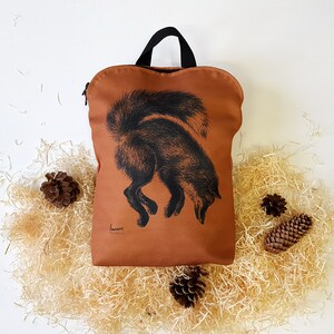 May include: A rust-colored backpack with a black handle and zipper. The front features a black illustration of a fox. The backpack is surrounded by wood shavings and pine cones.