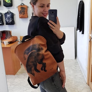 May include: A brown backpack with a black strap and a black fox illustration. The backpack is being worn by a person. Other bags with animal illustrations hang on the wall behind the person.