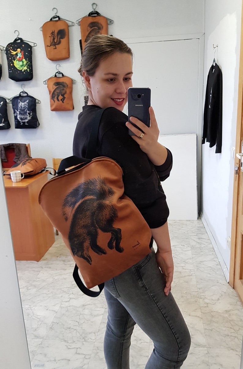 May include: A brown backpack with a black strap and a black fox illustration. The backpack is being worn by a person. Other bags with animal illustrations hang on the wall behind the person.