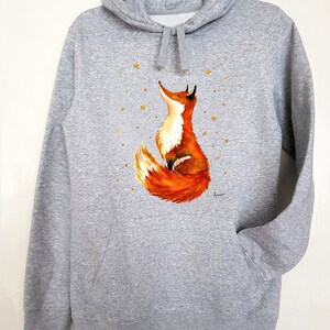 Puede incluir: Una sudadera con capucha gris con un bolsillo en la parte delantera. La sudadera presenta una ilustraci&oacute;n pintada a mano de un zorro rojo mirando hacia arriba a una dispersi&oacute;n de estrellas doradas.