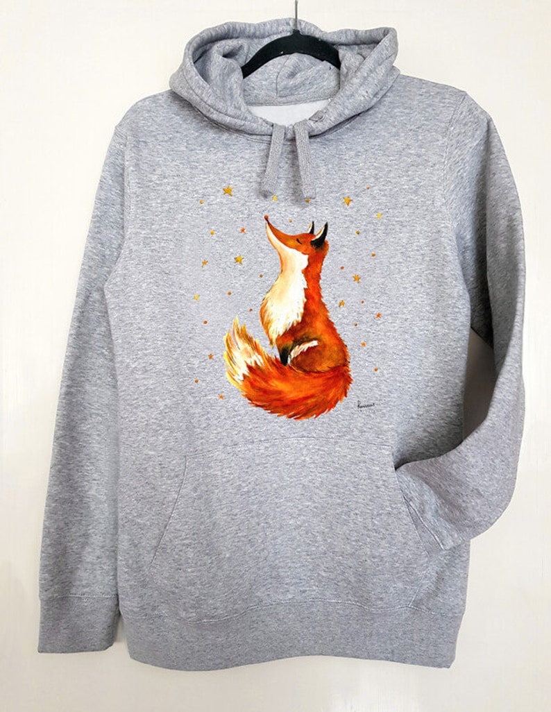 Puede incluir: Una sudadera con capucha gris con un bolsillo en la parte delantera. La sudadera presenta una ilustraci&oacute;n pintada a mano de un zorro rojo mirando hacia arriba a una dispersi&oacute;n de estrellas doradas.
