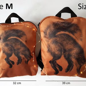 May include: Two orange backpacks with black handles and a black fox illustration. The size M backpack is 40 cm tall and 32 cm wide. The size L backpack is 44 cm tall and 39 cm wide.