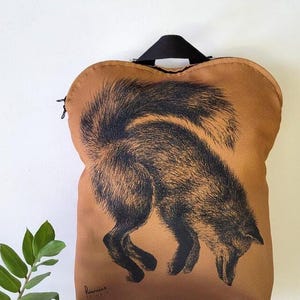 May include: A brown tote bag featuring a black illustration of a fox. The fox is depicted in a downward pose, with its head lowered and tail raised. The bag has a black handle and a zipper closure.