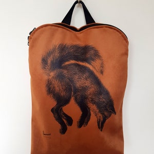 May include: A rust-colored backpack with a black fox illustration. The fox is depicted in a downward pose, with its tail arched. The bag has a black handle and zipper, and the brand name is printed at the bottom.