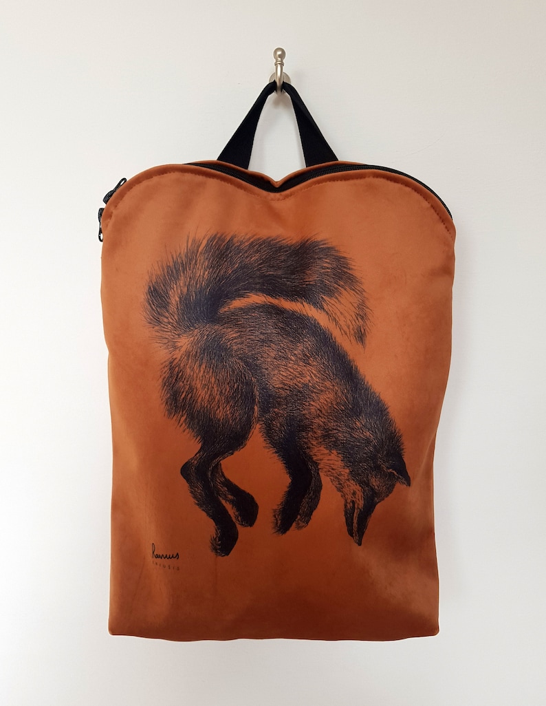 May include: A rust-colored backpack with a black fox illustration. The fox is depicted in a downward pose, with its tail arched. The bag has a black handle and zipper, and the brand name is printed at the bottom.
