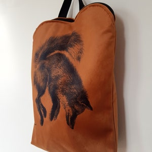 May include: A burnt orange tote bag with a black fox illustration. The bag has a black handle and zipper. The fox is depicted in a detailed, vintage style, with its head down and tail up. The bag is hanging on a silver hook.