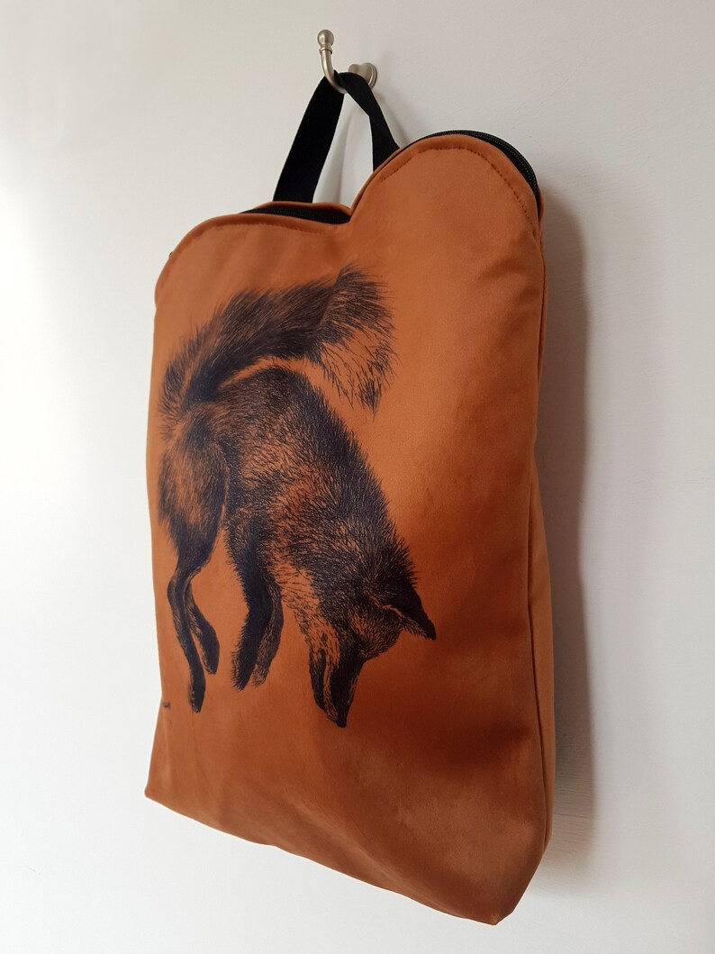 May include: A burnt orange tote bag with a black fox illustration. The bag has a black handle and zipper. The fox is depicted in a detailed, vintage style, with its head down and tail up. The bag is hanging on a silver hook.