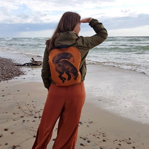 May include: An orange backpack with a black fox illustration. The backpack is worn over a green jacket. The person is standing on a sandy beach, looking out at the ocean. The person is wearing orange pants.