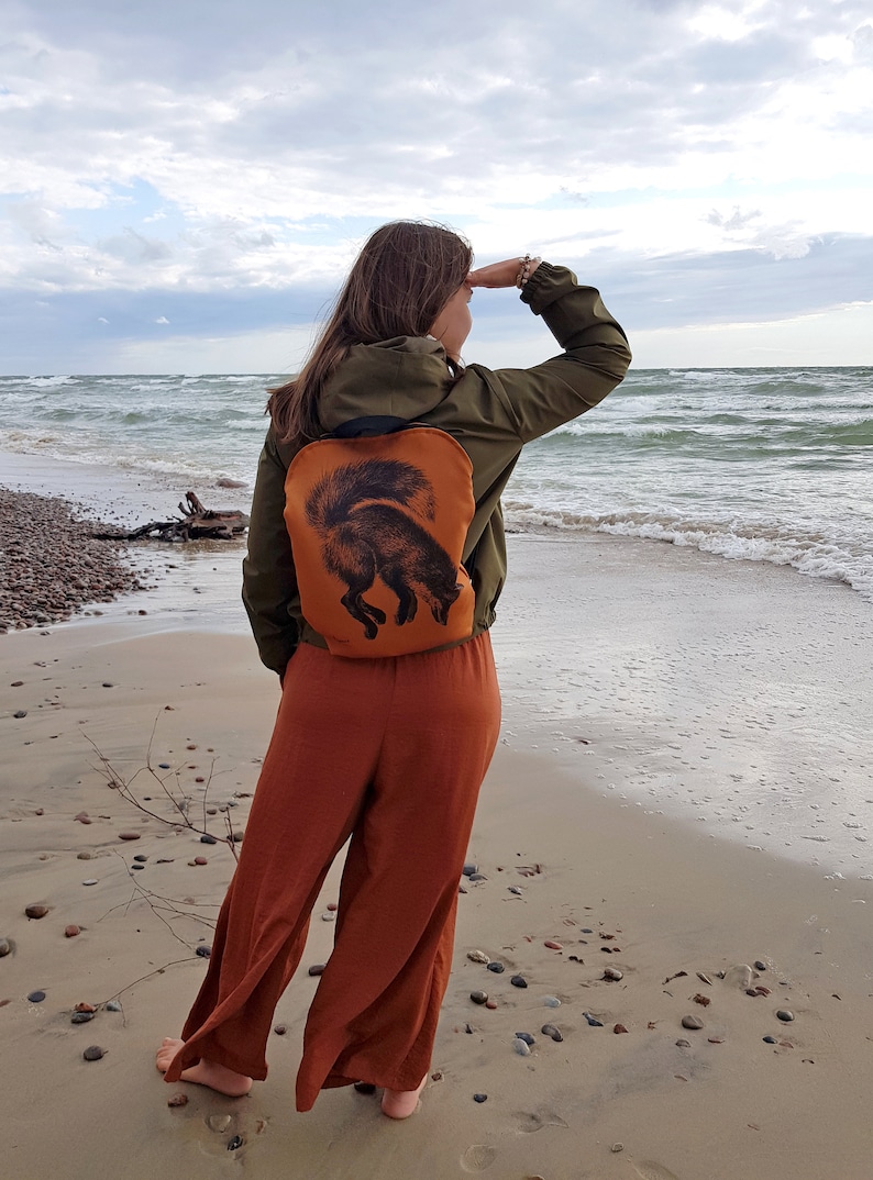 May include: An orange backpack with a black fox illustration. The backpack is worn over a green jacket. The person is standing on a sandy beach, looking out at the ocean. The person is wearing orange pants.