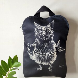 May include: Black backpack with a whimsical illustration of a cat holding a fish skeleton. The cat has large eyes and a fluffy appearance. The bag has a handle and a zipper closure.