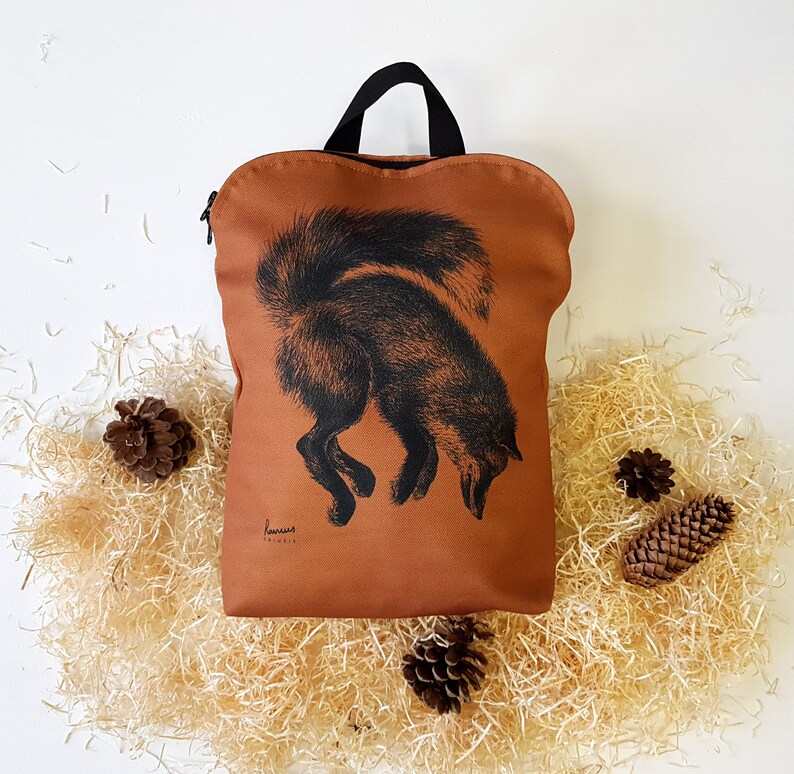 May include: A rust-colored backpack with a black handle and zipper. The front features a black illustration of a fox. The backpack is surrounded by wood shavings and pine cones.