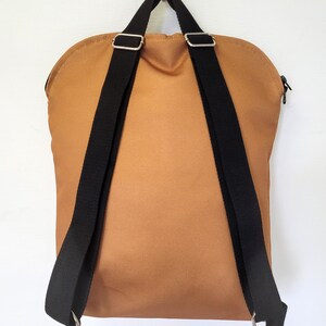 May include: A tan backpack with black adjustable straps and silver metal hardware. The backpack has a rounded top and a zipper closure. The straps are attached to the bottom with silver metal rings.