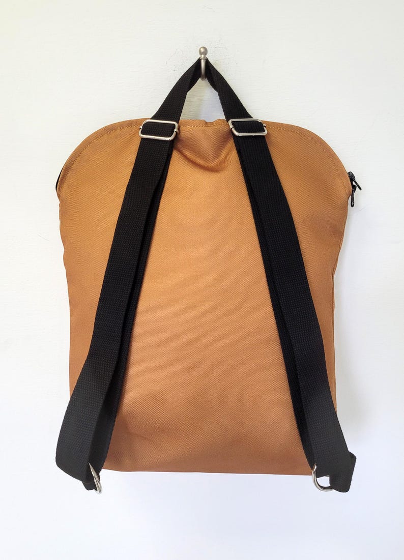 May include: A tan backpack with black adjustable straps and silver metal hardware. The backpack has a rounded top and a zipper closure. The straps are attached to the bottom with silver metal rings.