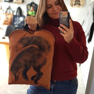 May include: A brown tote bag with a black fox illustration. The bag has a black handle and is held by a person. Other tote bags with various designs are hanging in the background.