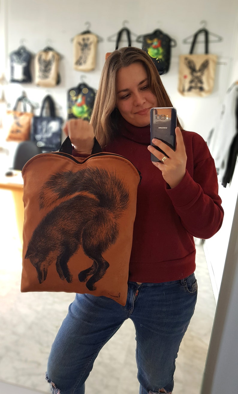 May include: A brown tote bag with a black fox illustration. The bag has a black handle and is held by a person. Other tote bags with various designs are hanging in the background.