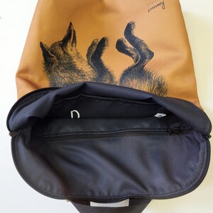 May include: A tan backpack with a black interior and a zipper closure. The backpack features a black and white illustration of a fox. The word "Rannus" is printed on the upper right corner of the backpack.