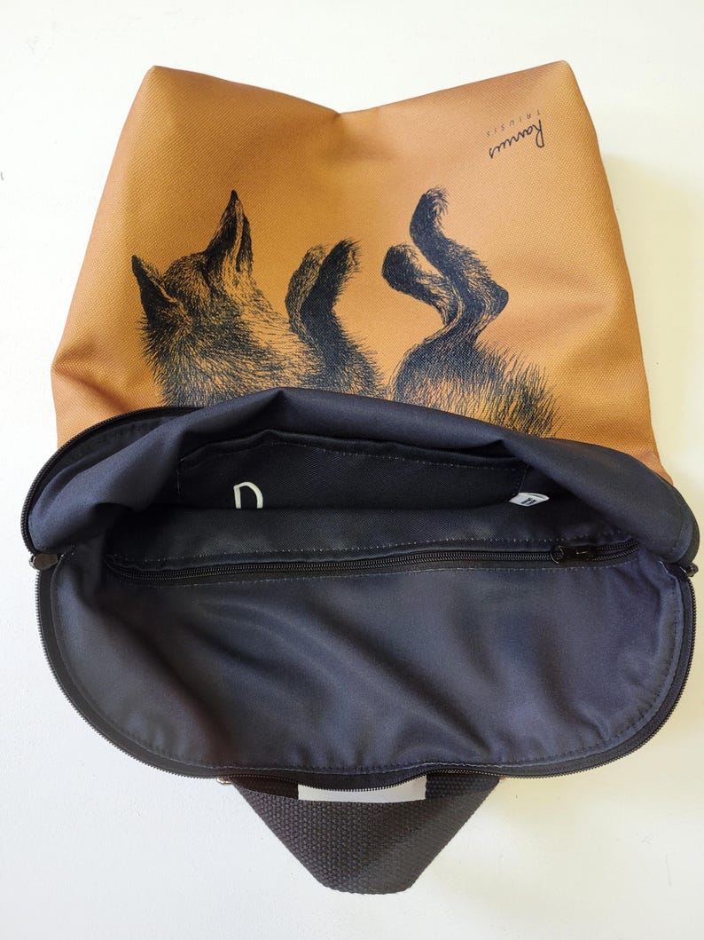 May include: A tan backpack with a black interior and a zipper closure. The backpack features a black and white illustration of a fox. The word "Rannus" is printed on the upper right corner of the backpack.