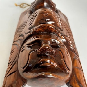 May include: A dark brown carved wooden wall mask featuring a prominent human face at the bottom and a smaller animal figure at the top. The wood has a polished finish with deep, detailed grooves and a small hanging loop attached to the top edge.
