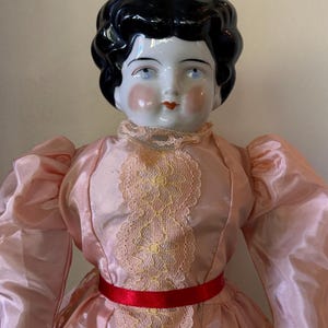 May include: A porcelain doll with a black, glossy hairstyle and a pale face with rosy cheeks and red lips. The doll wears a pink dress with lace detailing and a red ribbon belt. The doll's dress has long, puffy sleeves.