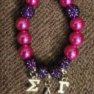 Puede incluir: Un brazalete de cuentas rosa y morado con dijes plateados que deletrean "ΣΑΓ".