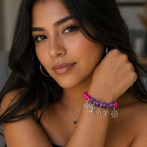 Könnte beinhalten: Ein Armband mit rosa und lila Perlen, silbernen Charms und einem Kronen-Charm. Das Armband wird am Handgelenk getragen. Eine goldene Halskette mit einem kleinen Anhänger ist ebenfalls sichtbar.