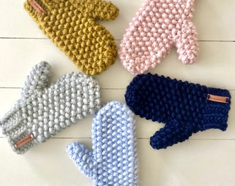 May include: Five pairs of knitted mittens in different colors: mustard yellow, light pink, light blue, gray, and navy blue. Each mitten has a small brown leather tag with the text "Woodland Knits" on it.