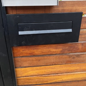 May include: A black metal letterbox with a silver slot for mail. The letterbox is mounted on a wooden surface.