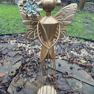 May include: A bronze-colored metal angel sculpture with a flower and wings. The angel has a round head, a halo, and a cage-like base. The wings and flower have a blue-green patina. The sculpture is outdoors on a wooden surface.
