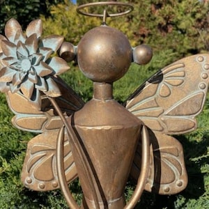 May include: Bronze angel garden statue with a halo, holding a flower. The angel has detailed butterfly wings and a stylized face. The statue is set against a backdrop of green foliage, adding a touch of whimsy to any outdoor space.