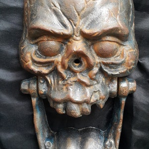 May include: A cast iron door knocker shaped like a skull with a wide open mouth. The skull is painted in shades of brown, blue, and gray, giving it a weathered look.