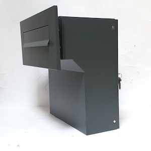 May include: A dark grey metal mailbox with a slot for mail and a keyhole. The mailbox has a built-in stand and is mounted to a wall.
