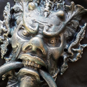 May include: A cast iron door knocker shaped like a grotesque face with a ring handle. The knocker is painted in a dark green and gold finish.