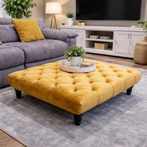 May include: A mustard yellow, square ottoman with a buttoned, tufted design. The ottoman sits on four short, black legs. It is placed on a grey rug in a living room. The ottoman's dimensions are approximately 90 cm x 90 cm.