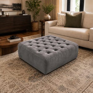 May include: A large, square, button-tufted ottoman in grey fabric. The ottoman is placed on a patterned rug. The room also features a sofa, coffee table, and decorative items. The ottoman is approximately 100 cm x 100 cm.