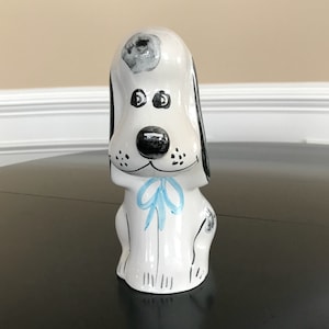 May include: A ceramic dog figurine with a white base, black and gray accents, and a light blue bow. The dog has large black eyes and a black nose. The figurine is a decorative item.
