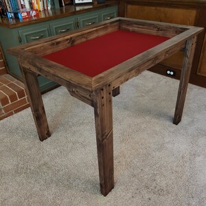 May include: A wooden game table with a red felt top and a dark brown stain. The table has a raised edge around the top and four sturdy legs.