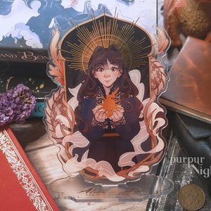 Puede incluir: Un soporte de acrílico transparente con un diseño en negro y dorado que presenta a una mujer con cabello largo y oscuro sosteniendo una llama dorada. El soporte tiene un fondo blanco y el texto "Alina Starkov" está impreso en la parte inferior.