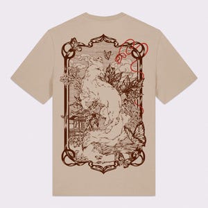 May include: Beige t-shirt with a detailed illustration of a white fox in a Japanese-inspired setting. The design includes a pagoda, butterflies, and foliage within a decorative frame. Red accents contrast the brown and beige colour scheme.